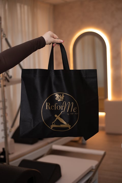 Person holding a black tote bag with ReforME gold logo in front of a glowing arch mirror